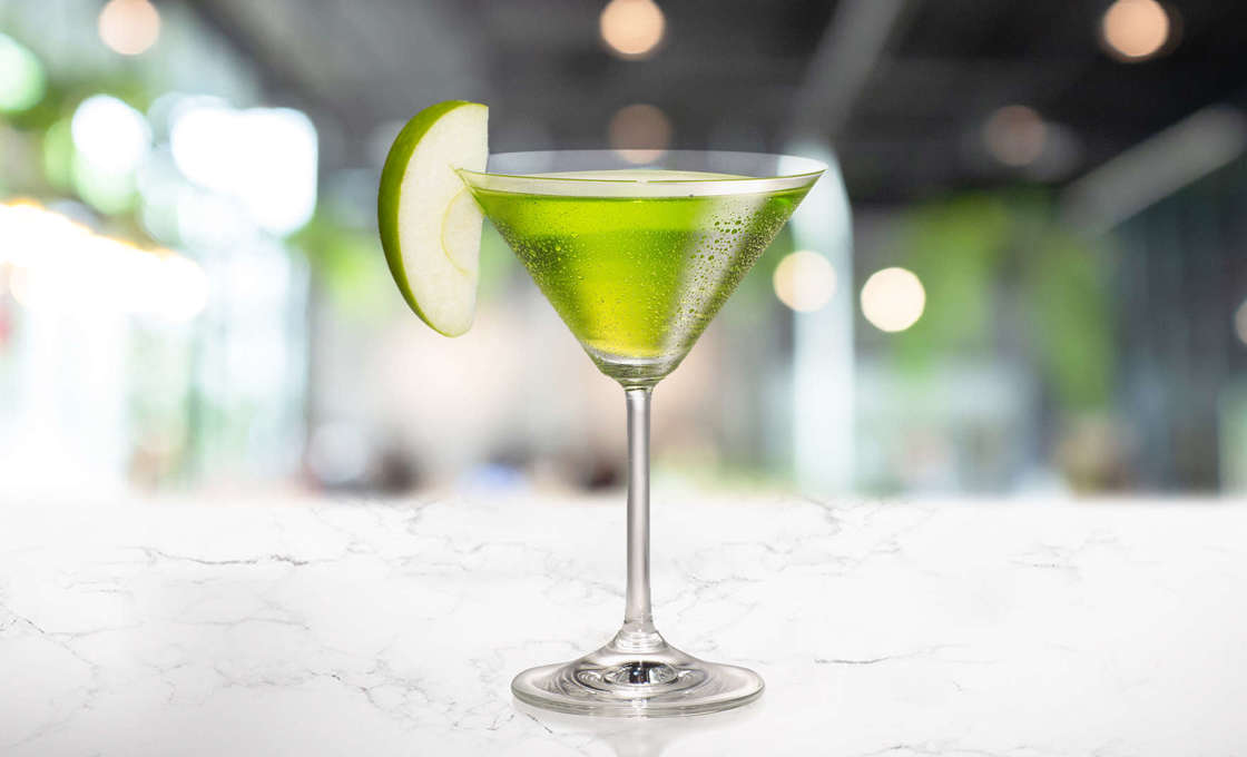 A glass of Appletini with a sliced apple on a white background