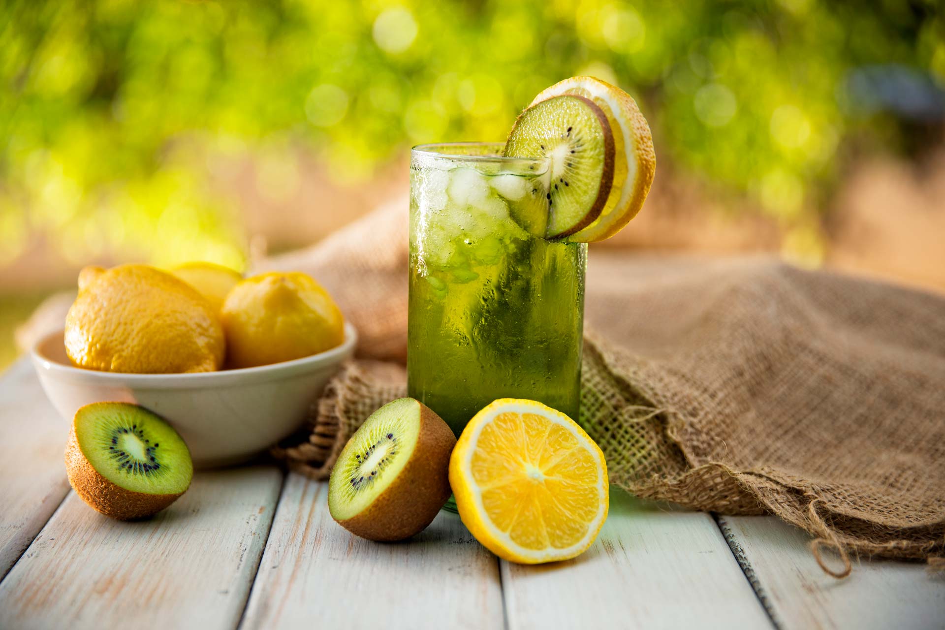 A glass of kiwi lime lemonade with ice on a table with lemons, limes and kiwis